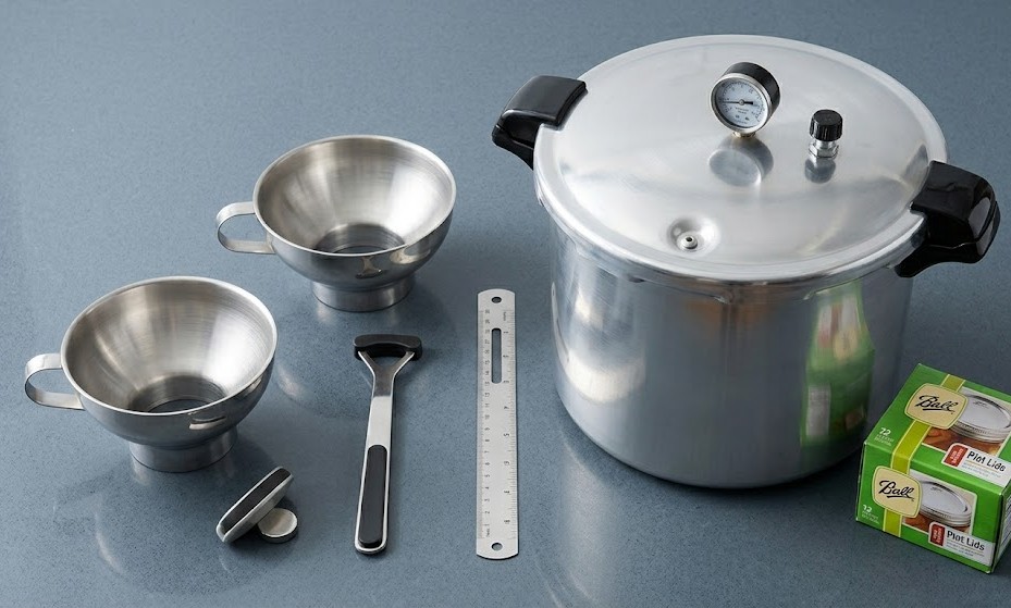 A flat lay arrangement of a pressure canner, funnel, headspace tool, and lids for preserving ranchero sauce.